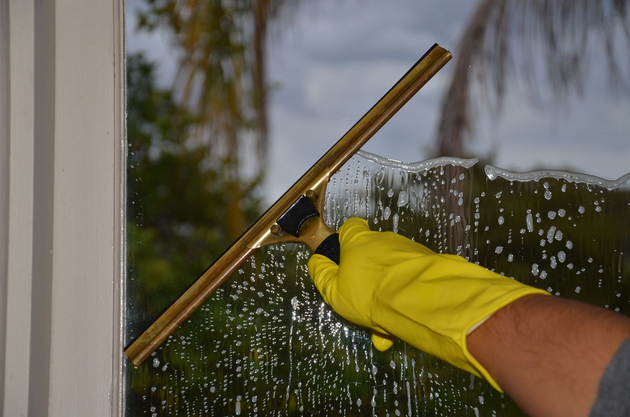 who-we-are Close-up of hand cleaning window with squeegee outdoors using yellow gloves.