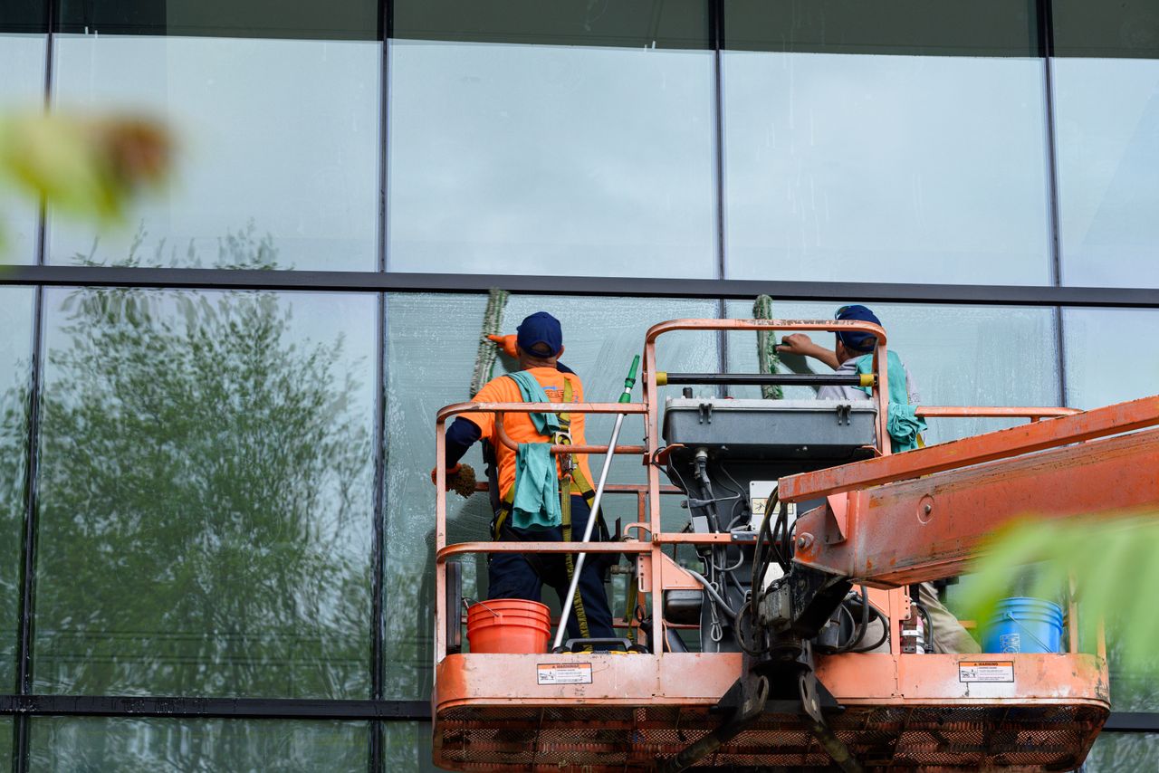 services-01 Two workers cleaning windows on a glass building facade using a lift platform, enhancing building aesthetics.