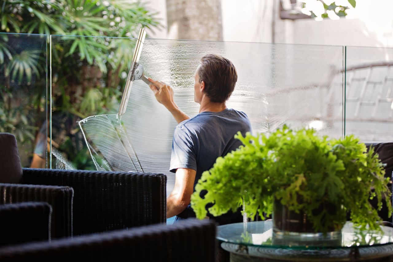 services-03 A man cleaning a glass barrier outdoors with a squeegee, surrounded by greenery.